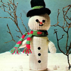 May include: A white knitted snowman with a black top hat, green and red scarf, and black button eyes. The snowman is standing in a snowy scene with bare branches.
