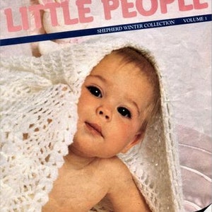 Puede incluir: Un bebé con una manta de ganchillo blanca, mirando a la cámara. La manta tiene un delicado patrón de encaje. La imagen es de la colección de invierno "Shepherd Little People".