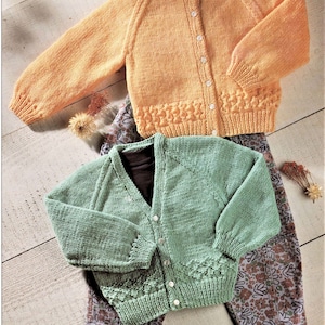 May include: Two knitted cardigans for children. One is a light green cardigan with a V-neck and button closure. The other is a light orange cardigan with a round neck and button closure.