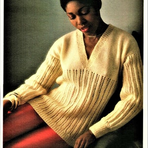 Puede incluir: Una mujer con un suéter de punto color crema con cuello en V y pantalones rojos. Está sentada y mirando un libro.