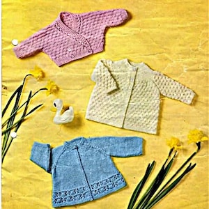 May include: Three knitted baby cardigans in pink, white and blue. The cardigans are laid out on a yellow background with flowers and a rubber duck.