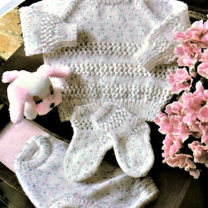 Può includere: Un maglione di lana bianco per neonato con un motivo a macchie rosa e bianco. Il maglione ha un collo rotondo e maniche lunghe. Ci sono anche pantaloni e guanti di lana abbinati per neonato.