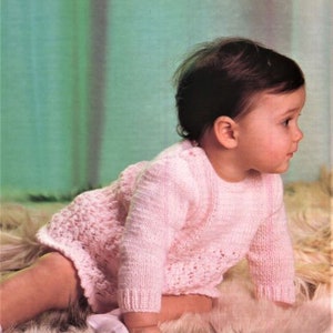 May include: A pink knitted baby dress with a lacy pattern. The dress has long sleeves and a round neckline. The baby is wearing the dress and crawling on a white furry surface.