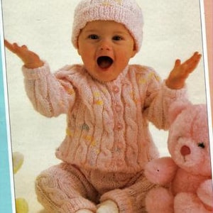 May include: A pink knitted baby outfit with a cable knit cardigan and matching trousers. The outfit is worn by a baby wearing a pink knitted hat. A pink teddy bear is sitting next to the baby.