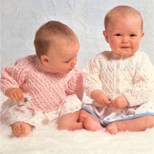 Può includere: Due bambini con maglioni di lana, uno rosa e uno bianco, seduti su una superficie bianca e morbida. I bambini indossano gonne bianche con bordi in pizzo. I maglioni presentano motivi a maglia complessi.