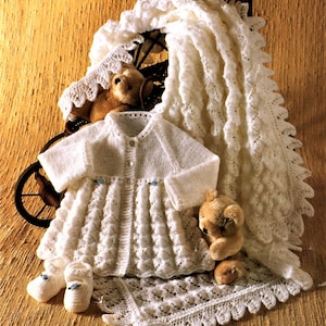 May include: A white knitted baby outfit set including a cardigan, dress, booties, and a blanket. The set is decorated with delicate lace trim and features a button closure on the cardigan.