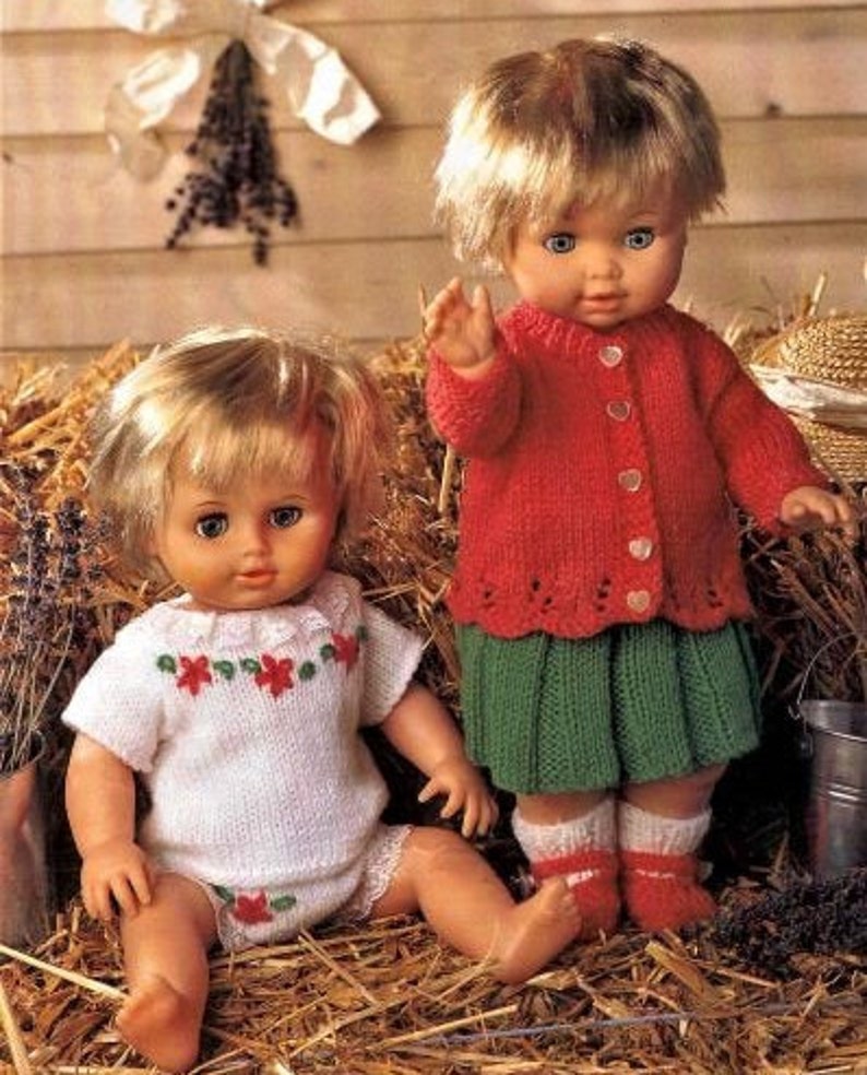 May include: Two dolls wearing knitted outfits. The doll on the left is wearing a white knitted dress with red and green floral embroidery. The doll on the right is wearing a red knitted cardigan with a green skirt.