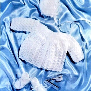 May include: A white crocheted baby outfit set, including a cardigan, bonnet, and booties. The set is made with 4 ply knit yarn and is displayed on a blue satin background.