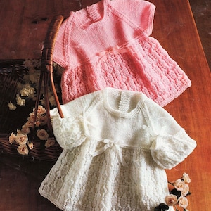 May include: Two knitted baby dresses, one white and one pink, with a lace pattern and a bow at the waist. The dresses are laid out on a wooden surface.