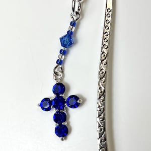 Könnte beinhalten: Silberner Lesezeichen mit einem Anhänger in Form eines blauen Kreuzes. Das Kreuz besteht aus runden, blauen Edelsteinen. Eine Kette aus blauen Perlen und ein blauer Kristall verbinden das Kreuz mit dem Haken des Lesezeichens. Das Lesezeichen hat ein dekoratives, geätztes Design.