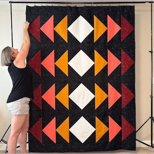 May include: A black, white, orange, and pink geometric quilt hanging on a stand. The quilt is made up of triangles and squares.