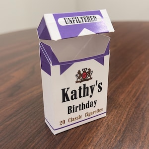 May include: A white and purple rectangular paper box with an open top lid. The front features the text Kathy's Birthday, 20 Classic Cigarettes, and a crest logo. The top flap displays the word UNFILTERED in black text on a white background.