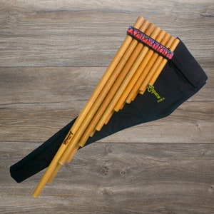 Puede incluir: Una flauta de pan de madera con una funda de transporte negra y una correa colorida. La flauta de pan está hecha de bambú natural y tiene una etiqueta verde que dice "Sopeca".