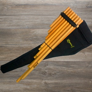 Puede incluir: Una flauta de pan de madera con una funda de transporte negra. La flauta de pan está hecha de bambú natural y tiene un color marrón. La funda tiene un exterior de tela negra y un logotipo verde que dice "Syos" en ella.