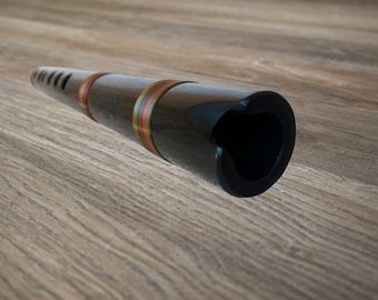 May include: A black wooden flute with a heart-shaped opening at the end. The flute has colorful bands of red, yellow, and green around the body. The instrument is set against a wooden background.