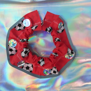 Op de afbeelding: Een rode stoffen halsband met een herhalend patroon van zwart-witte voetballen. De halsband heeft een grijze reflecterende rand en is versierd met verschillende ronde, iriserende, holografische accenten op het oppervlak.