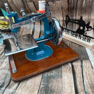 Peut inclure: Une machine à coudre bleu sarcelle de style vintage montée sur une base en bois rectangulaire. La machine présente une plaque à aiguille en métal argenté, une manivelle et une bobine de fil blanc sur le dessus. Un panneau noir porte le texte LIL SEW N SEW.