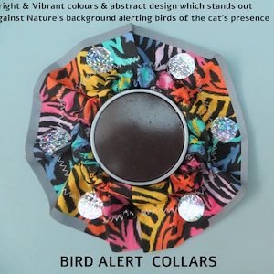 Könnte beinhalten: Ein Vogelwarnhalsband mit einem lebendigen, abstrakten Zebrastreifenmuster in Orange, Pink, Blau und Schwarz. Das Halsband hat einen zentralen dunklen Kreis und ist mit reflektierenden Akzenten verziert. Der Text "BIRD ALERT COLLARS" befindet sich unten.