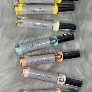 May include: An assortment of perfume oil roll-on bottles in various colors, including yellow, blue, and pink. Each bottle has a silver cap and a label that reads "Milar Aromar" and "Perfume Oil Roll-On". The bottles are arranged on a white, fluffy surface.
