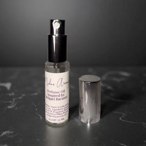 May include: A clear glass perfume oil bottle with a silver cap and black sprayer. The label reads "Mikas Aromas Perfume Oil Inspired by Bvlgari Baciama". The bottle is 0.5 oz / 15 ml.