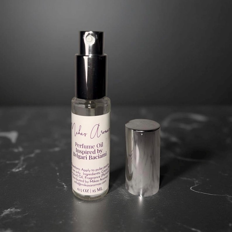 May include: A clear glass perfume oil bottle with a silver cap and black sprayer. The label reads "Mikas Aromas Perfume Oil Inspired by Bvlgari Baciama". The bottle is 0.5 oz / 15 ml.