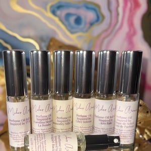 May include: Six perfume oil roll-on bottles with silver caps are displayed. Each bottle has a label with the brand name and the fragrance it is inspired by. The bottles are arranged on a reflective surface, with one bottle lying down.