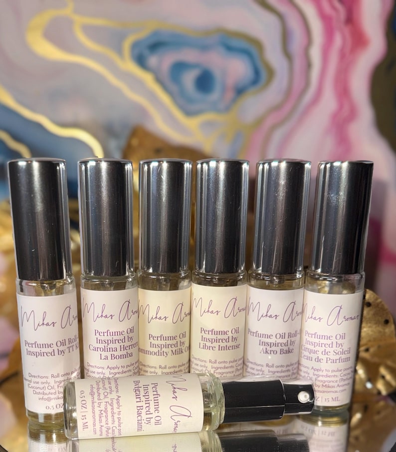 May include: Six perfume oil roll-on bottles with silver caps are displayed. Each bottle has a label with the brand name and the fragrance it is inspired by. The bottles are arranged on a reflective surface, with one bottle lying down.