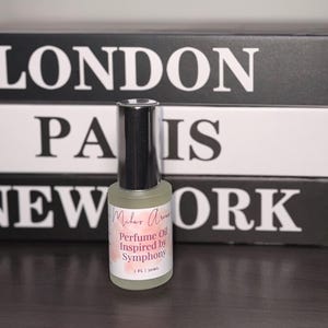 May include: A perfume oil bottle with a black cap and a label that reads "Milar Aroma Perfume Oil Inspired by Symphony". The bottle is clear, and the liquid inside is light yellow. The background features stacked books with the words "LONDON", "PARIS", and "NEW YORK".