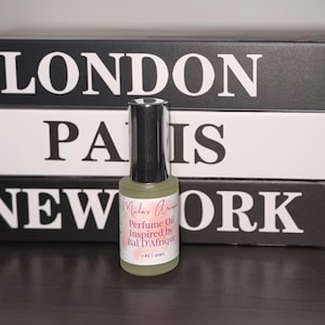 May include: A small perfume oil bottle with a black cap and a label that reads "Milar Aroma Perfume Oil Inspired by Bal D'Afrique." The bottle is set against a backdrop of black and white books with city names.
