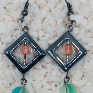 Peut inclure: Une paire de boucles d'oreilles pendantes avec un design géométrique. Les boucles d'oreilles sont en métal noir et présentent une forme carrée avec un plus petit carré à l'intérieur. Les boucles d'oreilles sont rehaussées d'une petite perle ronde orange et d'une plus grosse perle ronde bleu et verte.