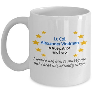May include: White ceramic coffee mug with a blue and yellow design. The mug features the text "Lt. Col. Alexander Vindman A true patriot and hero. I would ask him to marry me but I hear he's already taken."