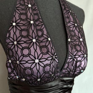 May include: A formal halter dress with a black satin waistband. The bodice features a black floral lace overlay with sparkling embellishments over a lavender lining. The dress is displayed on a black mannequin.