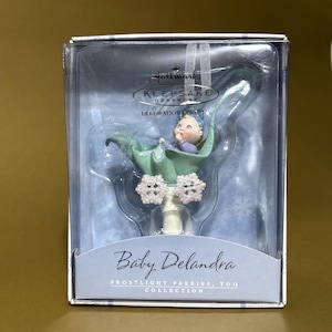 May include: A Hallmark Keepsake ornament featuring a baby fairy named Delandra from the Frostlight Faeries collection. The ornament is in a clear plastic box. The baby fairy is in a green leaf cradle with snowflake accents.