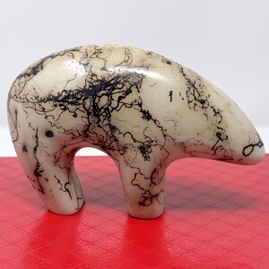 May include: A smooth, cream-colored ceramic bear figurine featuring a distinct black horsehair-style pattern of fine, intricate, web-like lines across its entire surface. The bear is depicted in a standing position with a rounded back and head.