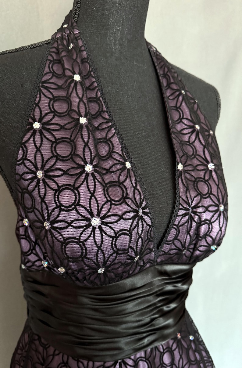 May include: A formal halter dress with a black satin waistband. The bodice features a black floral lace overlay with sparkling embellishments over a lavender lining. The dress is displayed on a black mannequin.