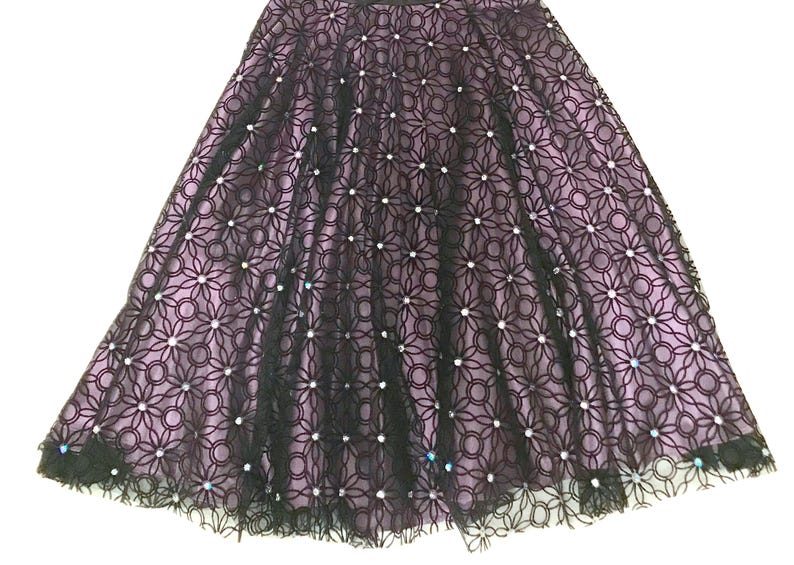 May include: A black lace skirt with a floral pattern and a lavender lining. The skirt is embellished with small, sparkling accents. The skirt has a flared silhouette, suitable for formal occasions.
