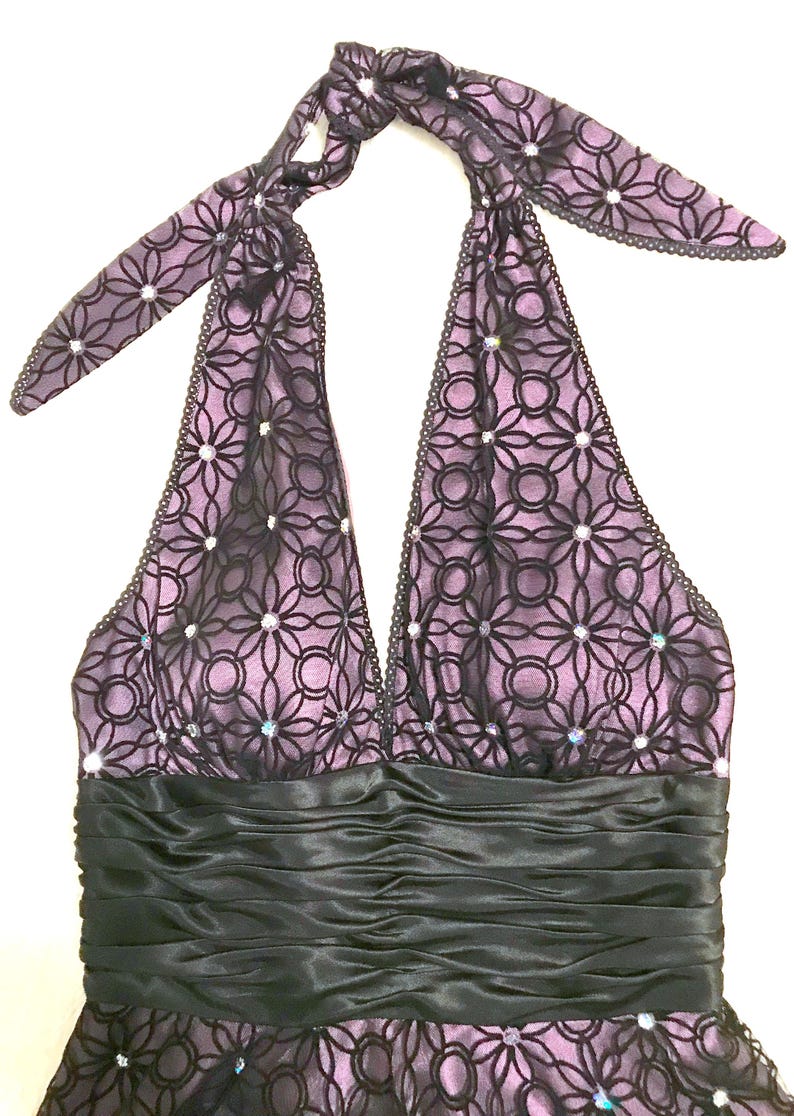 May include: A halter-style dress in shades of purple and black. The bodice features a floral lace overlay with sparkling accents. A black satin band defines the waist, and the halter straps are tied at the back of the neck. Formal wear.