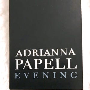 May include: A black rectangular tag with the text "ADRIANNA PAPELL EVENING" in white and gray lettering. The tag is set against a white, textured background, suggesting a fabric or textile.