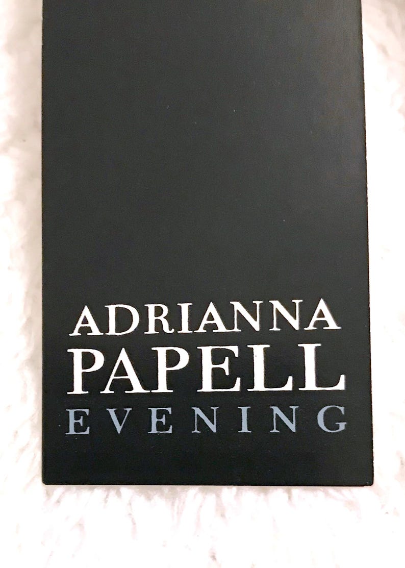 May include: A black rectangular tag with the text "ADRIANNA PAPELL EVENING" in white and gray lettering. The tag is set against a white, textured background, suggesting a fabric or textile.
