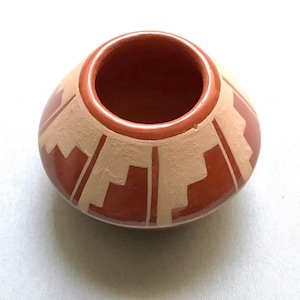 May include: A small, round, terracotta-colored pottery bowl with a wide opening. The bowl features a geometric design in a lighter tan color. The rim is a darker shade of terracotta. The bowl is likely handmade.
