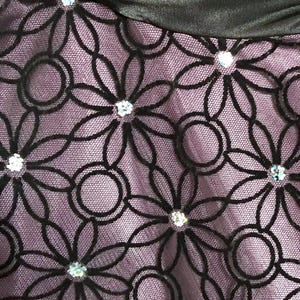 May include: Close-up of a fabric with a floral pattern. The design features black, stylized flowers with circular accents, set against a lavender background. Sparkling embellishments are placed at the center of each flower.