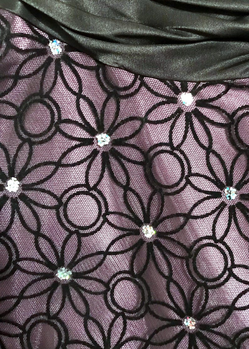 May include: Close-up of a fabric with a floral pattern. The design features black, stylized flowers with circular accents, set against a lavender background. Sparkling embellishments are placed at the center of each flower.