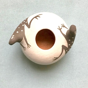 May include: A white ceramic ring-shaped object with a brown interior and two gray frog designs. The frogs have white polka dots and are positioned on opposite sides of the ring. The background is a light blue fabric.