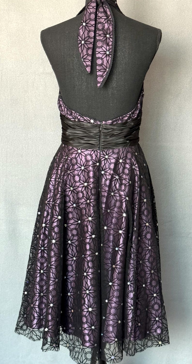 May include: A formal halter dress with a black bodice and a full skirt. The skirt features a floral lace overlay over a lavender fabric, embellished with small, sparkling accents. The dress has a black satin waistband and a matching halter tie.