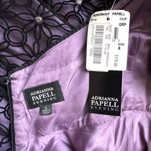 May include: A close-up of a lavender evening dress by Adrianna Papell. The dress features a black floral lace overlay with rhinestone accents. The interior lining is a smooth, satin-like fabric. A tag indicates the size is 6.