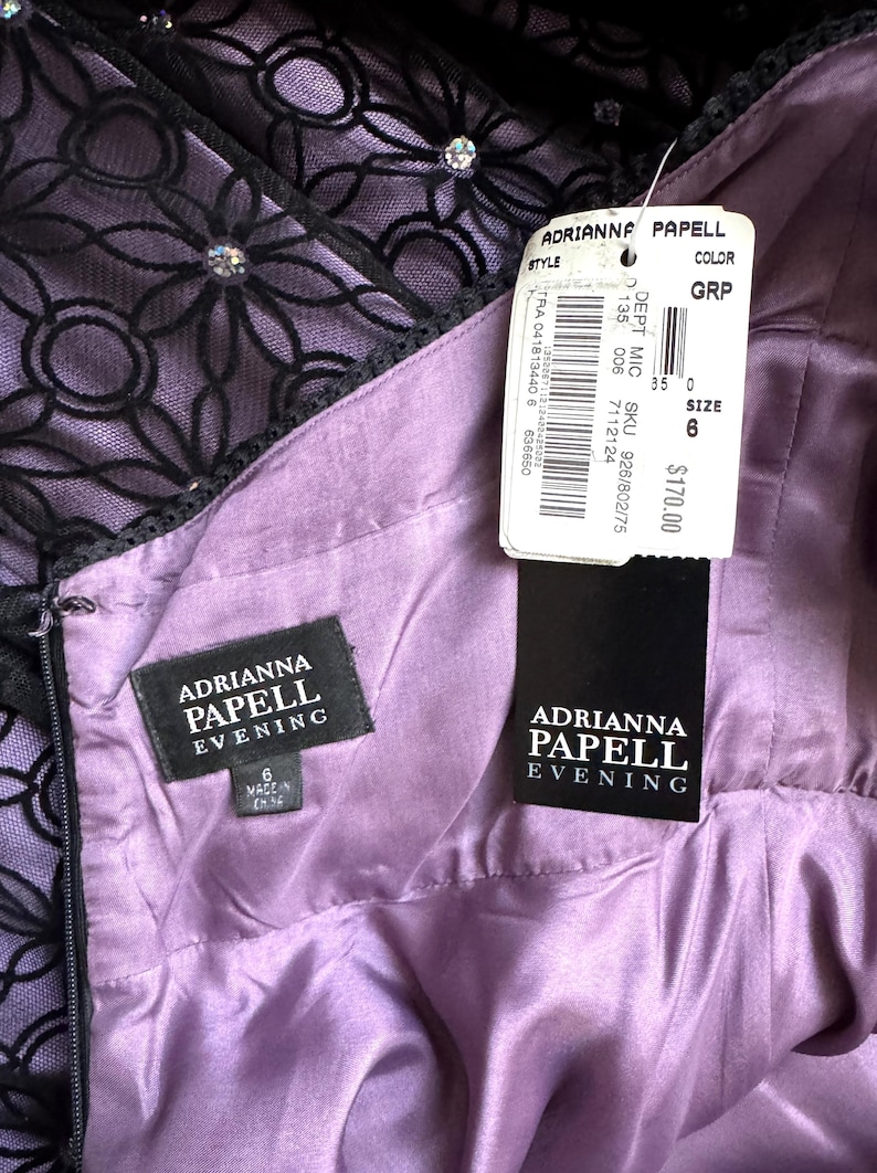 May include: A close-up of a lavender evening dress by Adrianna Papell. The dress features a black floral lace overlay with rhinestone accents. The interior lining is a smooth, satin-like fabric. A tag indicates the size is 6.