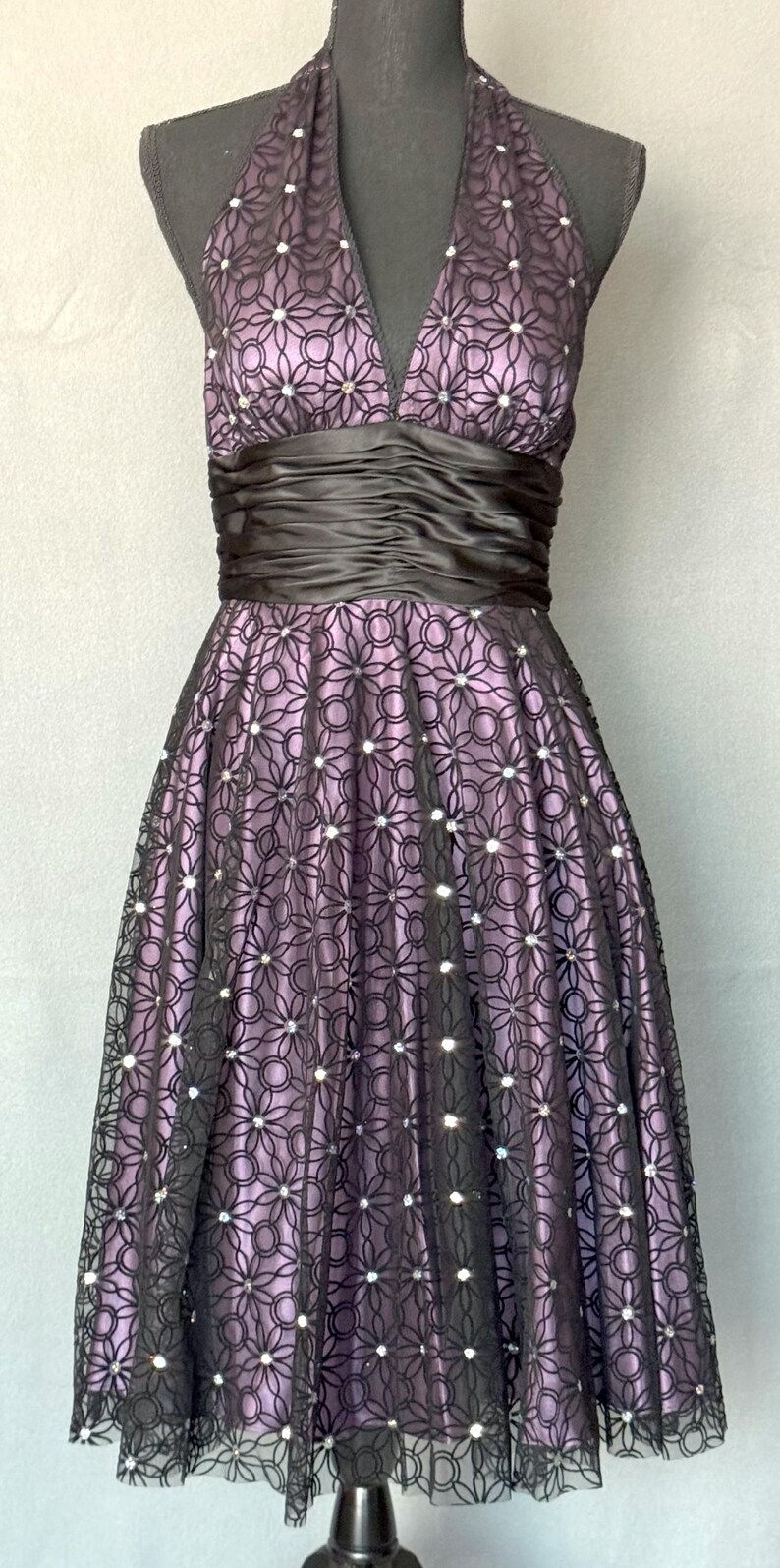 May include: A formal halter dress in shades of purple and black. The bodice features a V-neck and a ruched black satin band at the waist. The skirt is a full A-line, with a floral lace overlay and sparkling accents. The dress is displayed on a mannequin.