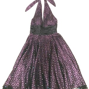 May include: A halter-neck dress in a formal style. The dress features a black lace overlay with a floral pattern over a lavender lining. A black satin ribbon defines the waist. The skirt flares out, and the hemline is trimmed with black lace.
