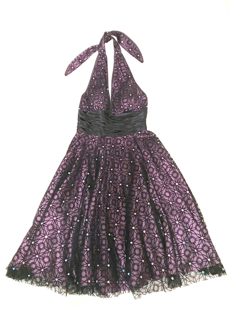 May include: A halter-neck dress in a formal style. The dress features a black lace overlay with a floral pattern over a lavender lining. A black satin ribbon defines the waist. The skirt flares out, and the hemline is trimmed with black lace.