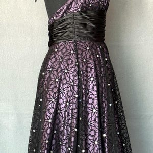 May include: A formal halter dress with a black floral lace overlay over a lavender lining. The dress features a black satin waistband and is adorned with small, sparkling embellishments.